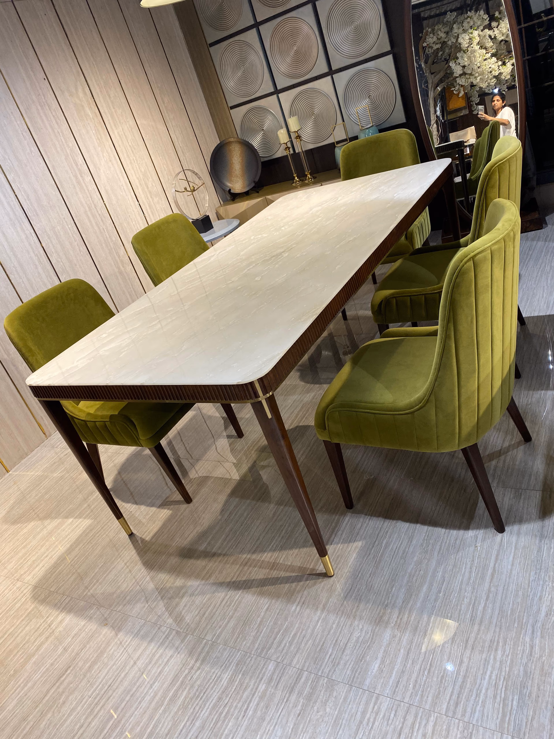 Angled view along the white marble dining table showing the fluted walnut apron, brass leg caps and the olive-green velvet chairs tucked underneath.