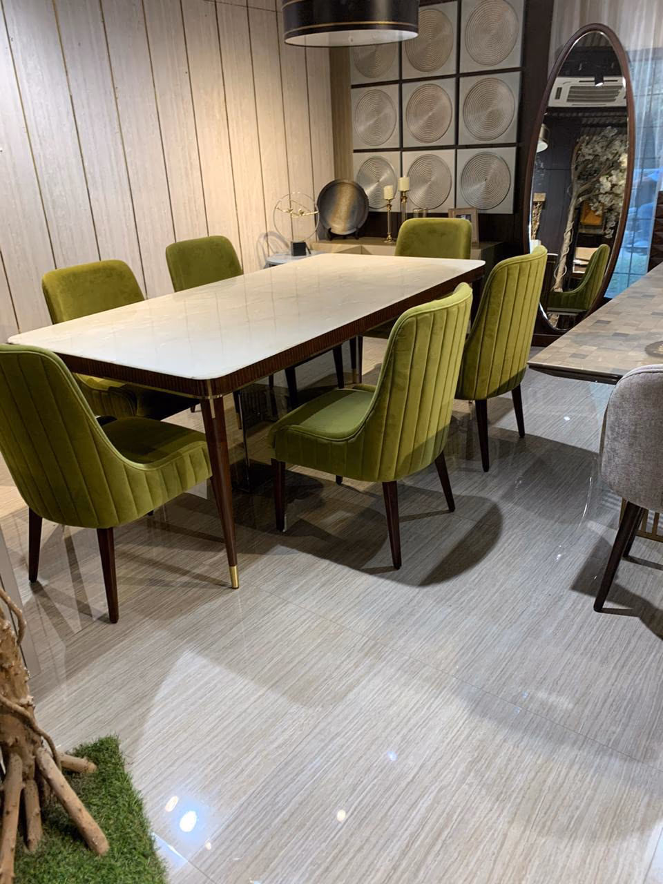 Full view of the marble-top dining table set with six olive-green velvet chairs in front of a tall oval mirror and ribbed wall panelling.