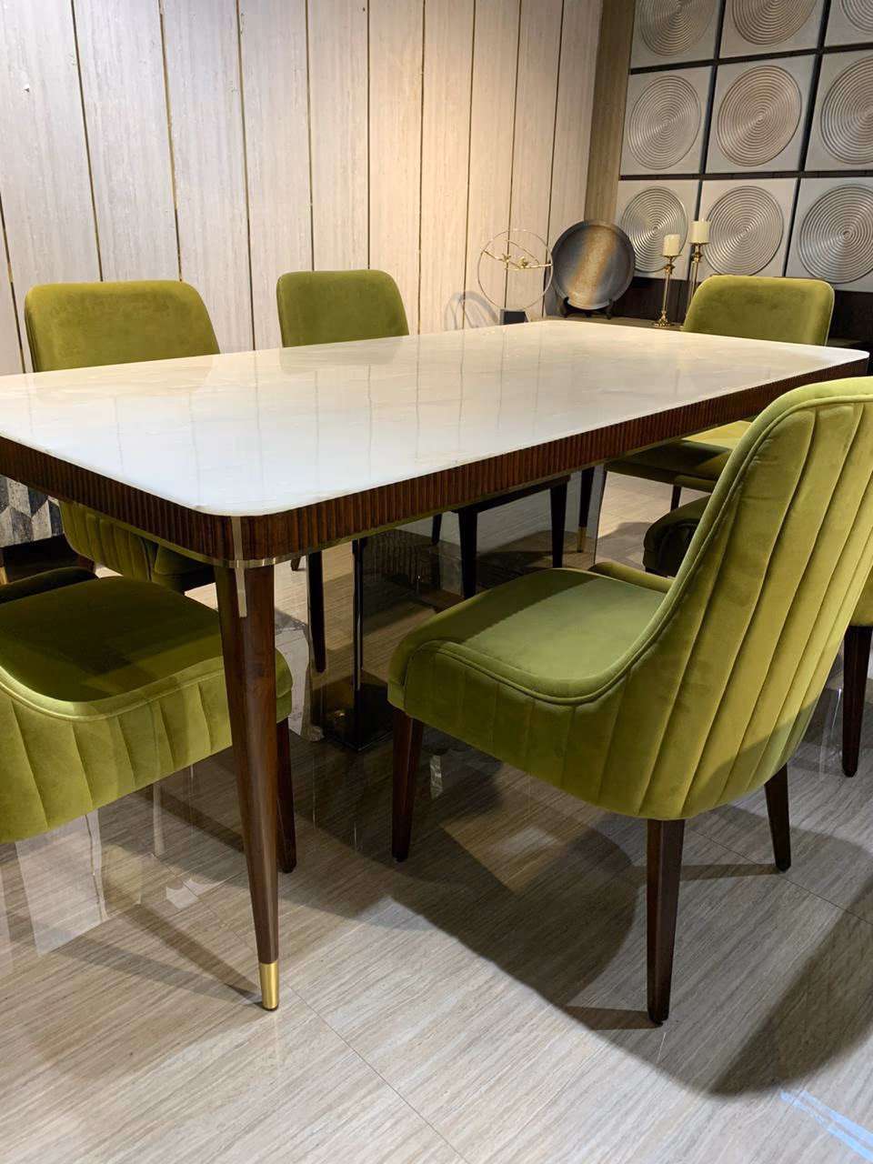 Close view of the white marble dining table corner with olive-green velvet chairs pulled in around it and a tapered brass-capped leg in the foreground.