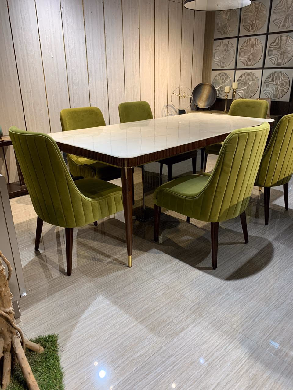 Rectangular dining table with a white marble top and fluted walnut apron on tapered brass-capped legs, surrounded by six olive-green channel-tufted velvet chairs.