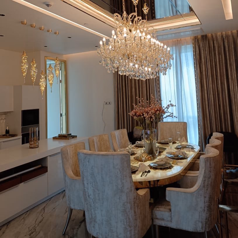Eight-seat dining set with a golden onyx table top and taupe crushed-velvet armchairs on slim chrome legs, lit by a tiered crystal chandelier beside a white kitchen island.