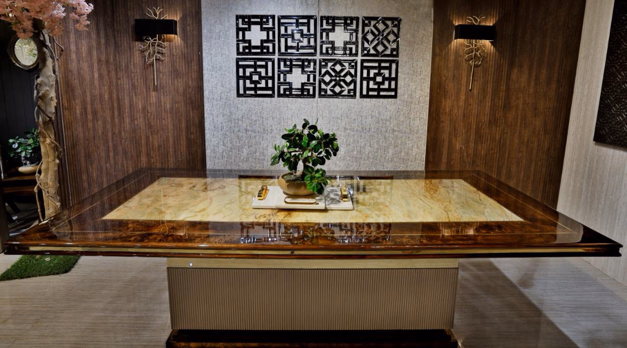 Overhead-angled view of the dining table showing the full honey onyx slab framed in glossy burl wood, with a bonsai on a marble tray at the centre.