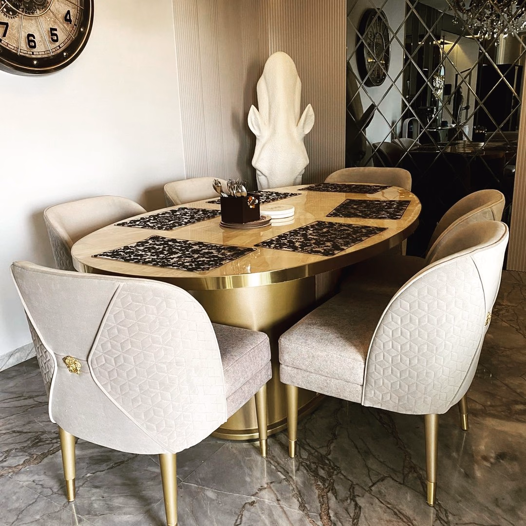 Oval dining table with a book-matched inlay top and a polished brass drum pedestal on fluted legs, surrounded by cream quilted dining chairs with gold medallion backs.