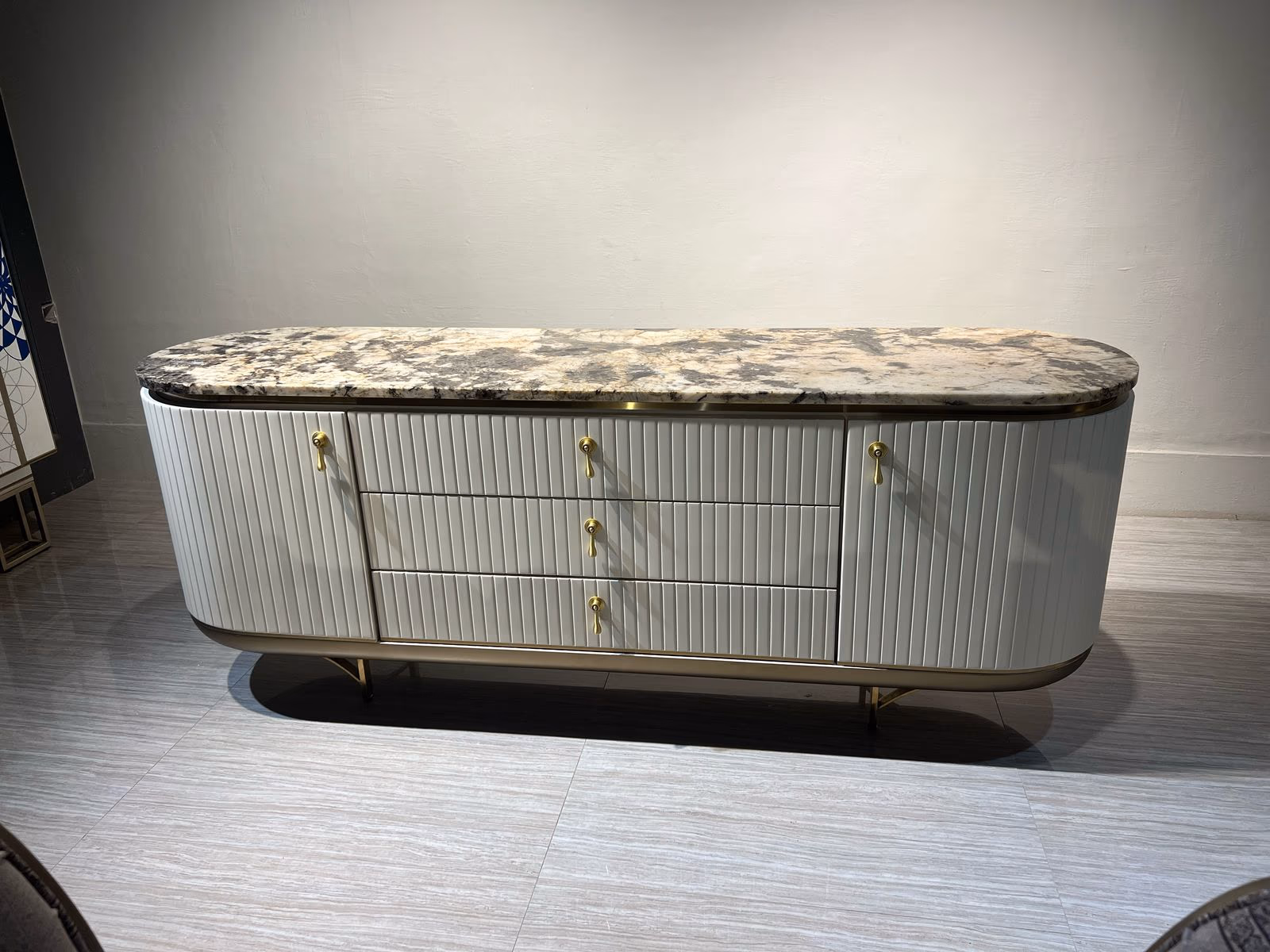 Rounded white fluted sideboard with three center drawers, two side doors, brass hardware, and a grey-gold marble top.