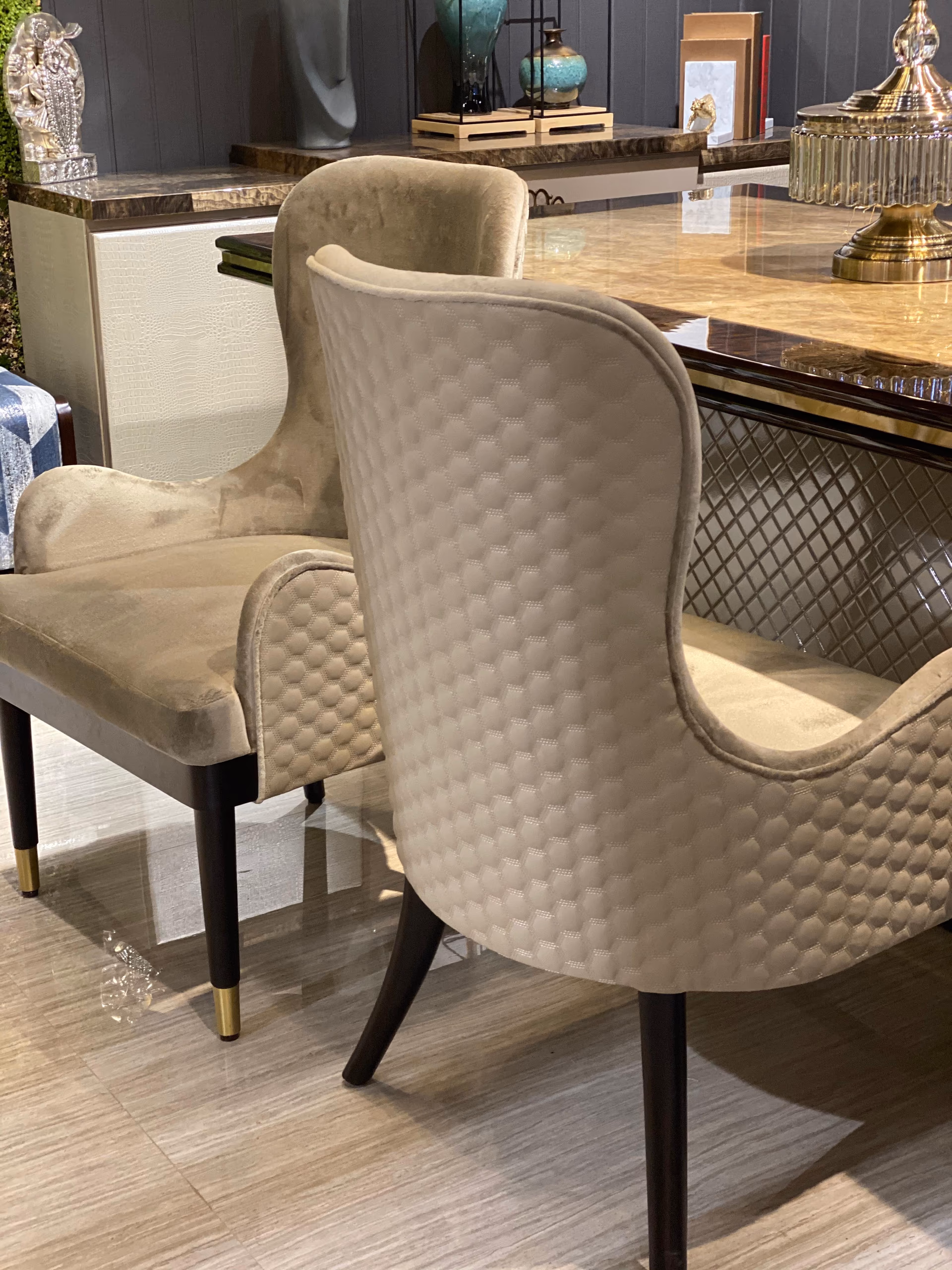 Two taupe velvet dining chairs at an onyx table, one a carver with arms, showing a curved wingback shape and honeycomb-quilted outer backs on tapered wood legs.