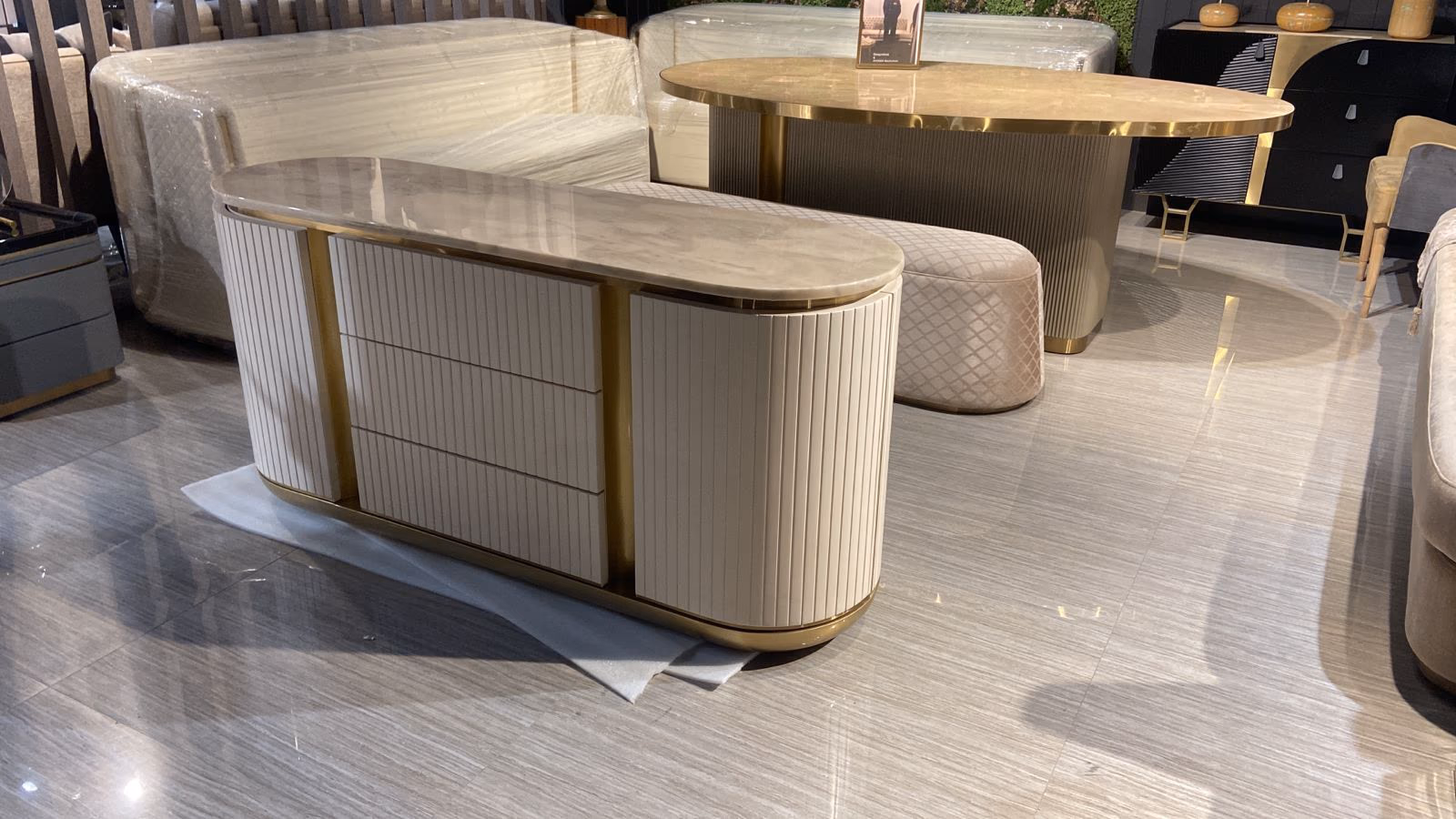 Showroom vignette of a cream fluted sideboard in the foreground and an oval gold-top dining table with a quilted ottoman behind it.