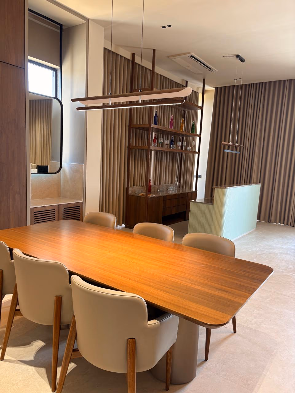 Rich walnut-grain dining table with six cream barrel chairs beside a dark wood bar shelf and linear pendant lights.