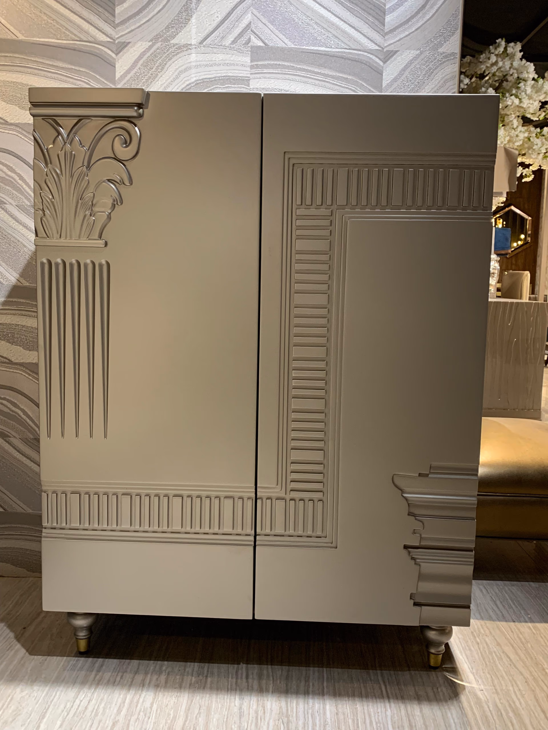 Tall taupe two-door cabinet carved with an Art Deco palm capital, fluted pilaster and stepped dentil mouldings, on turned brass-capped feet.