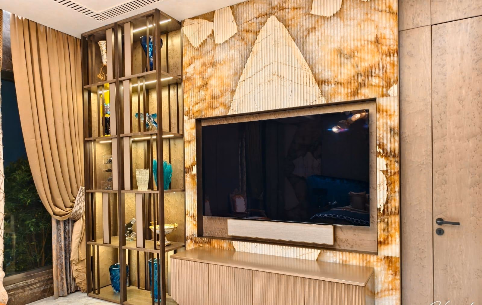 Floor-to-ceiling brass-framed open display shelving with lit glass niches, standing beside a backlit amber onyx television wall and a fluted media console.