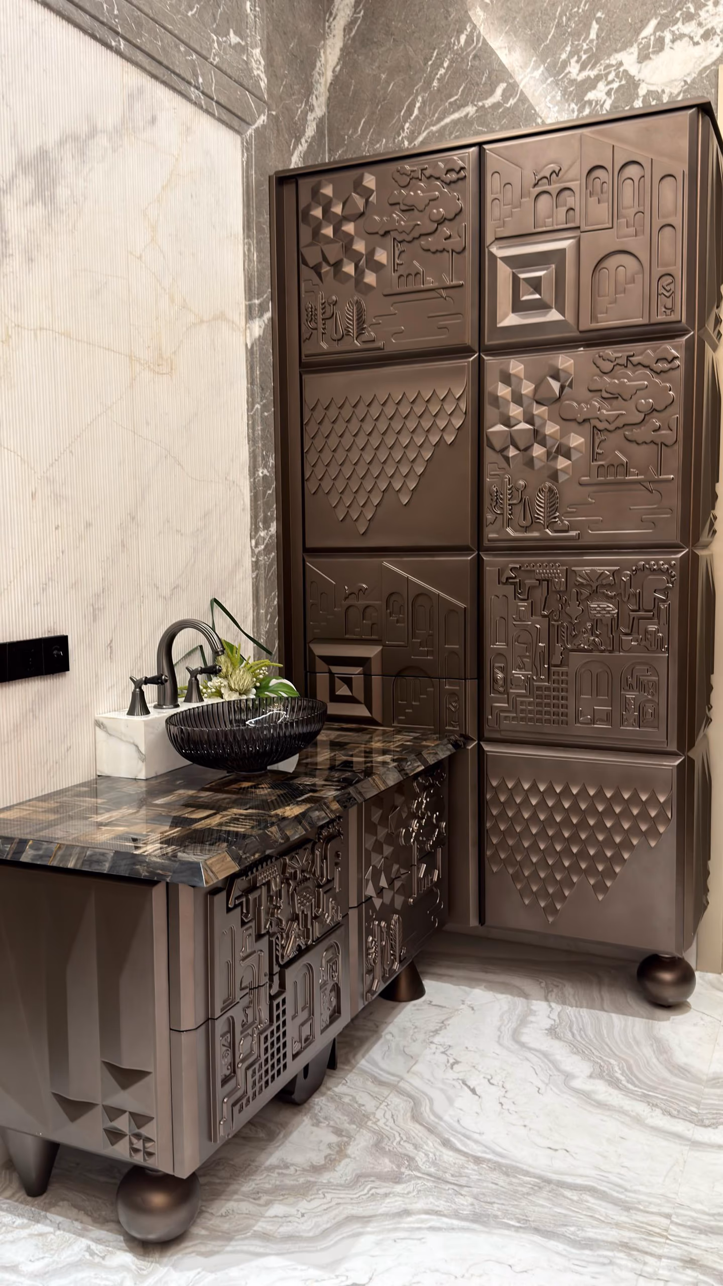 The relief-panel furniture in a warm bronze finish, a tall sculpted cabinet beside a matching relief vanity with a dark glass bowl, in a white marble powder room.