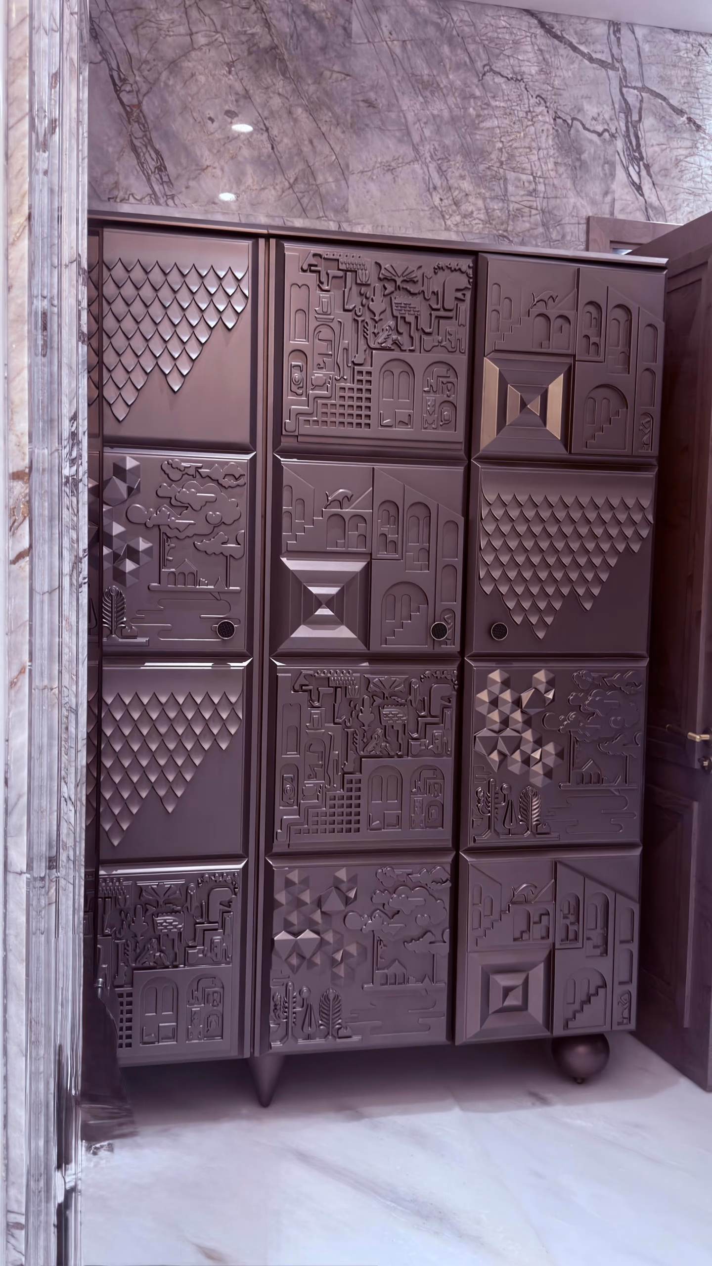 Tall pewter-toned cabinet whose doors are covered in deep sculpted relief panels of townscapes, trees, arches and pyramids, standing on a marble floor.
