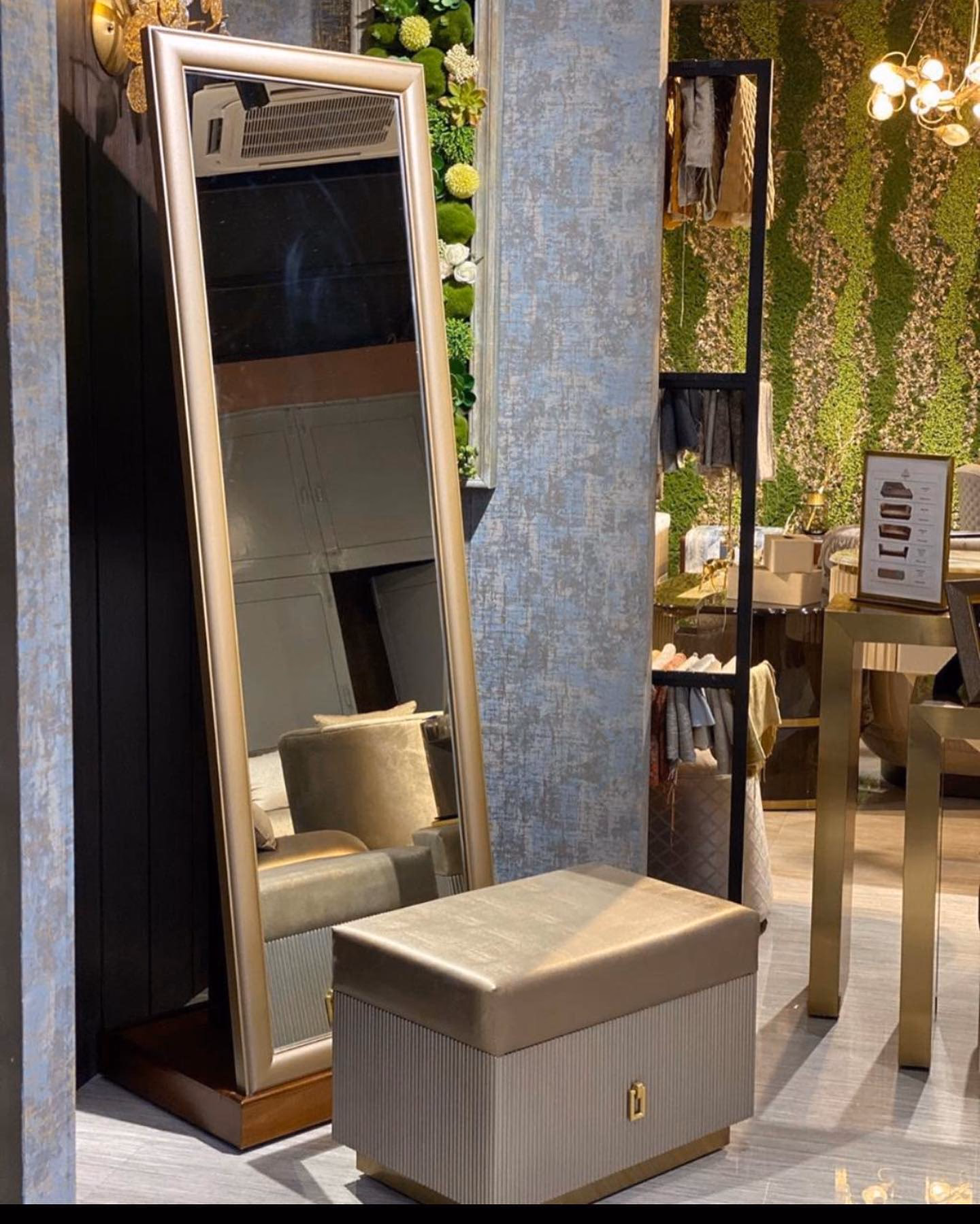 Tall champagne-framed floor mirror on a dark wood base standing beside a fluted grey storage ottoman with a gold leather lid, a brass turn-catch and a brass plinth.
