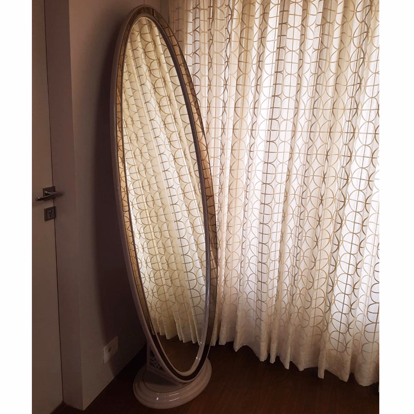 Large oval cheval floor mirror in a plain champagne frame on a round moulded pedestal foot, tilted against a bedroom wall beside sheer patterned curtains.