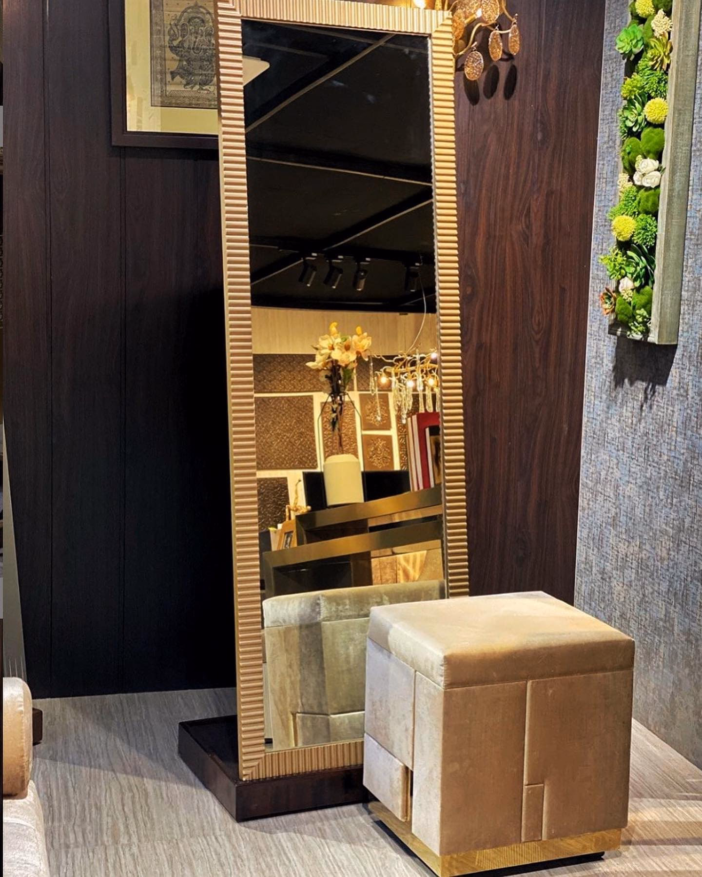 Full-length rectangular mirror in a wide hand-fluted gold frame standing on a dark wood plinth, with a tan suede panelled cube pouf on a brass base beside it.