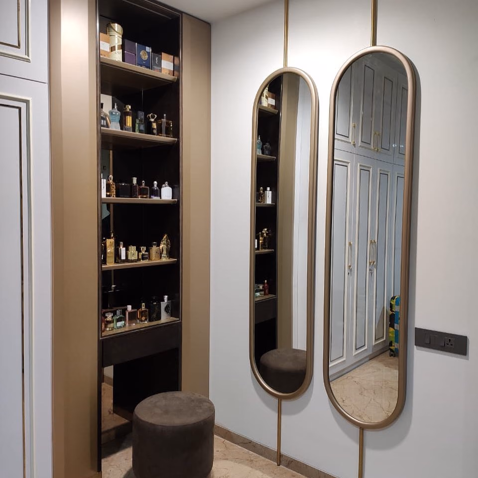 Pair of tall capsule-shaped mirrors in slim brushed gold frames mounted on a vertical gold rail, beside a lit perfume shelf niche and a brown velvet round pouf in a dressing room.
