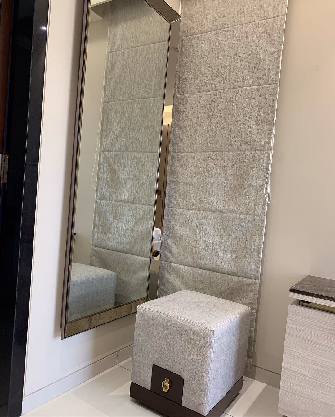 Tall narrow wall mirror in a slim bronze metal frame mounted flush to a dressing wall, with a grey linen cube stool banded in dark wood and a brass turn-catch below it.