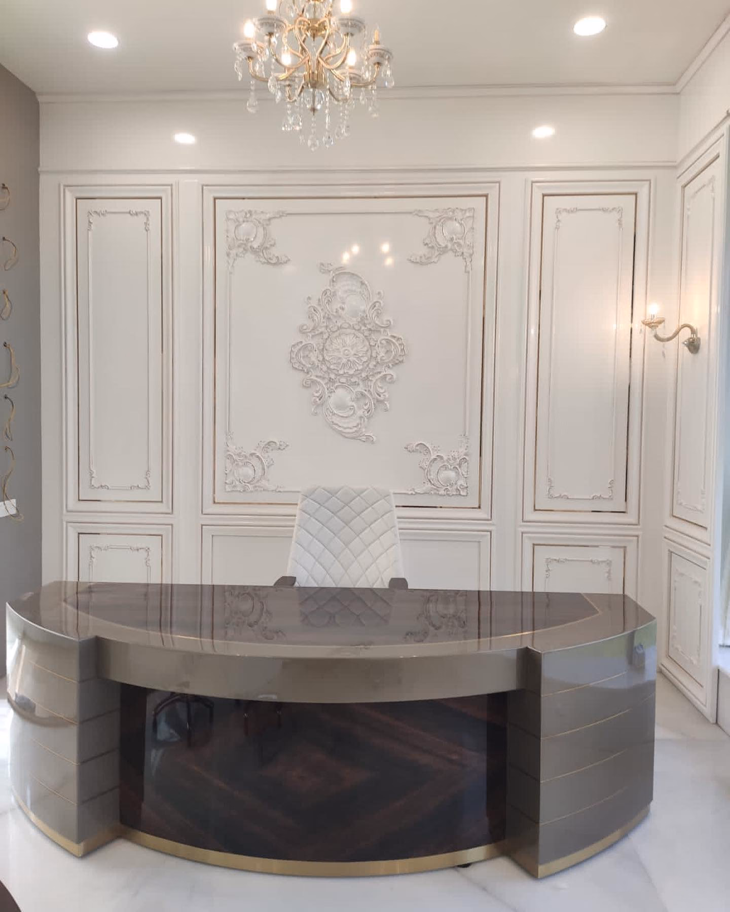 D-shaped director's desk with a grey stone top, champagne lacquer end pods, a burl-wood modesty panel and a brass plinth, set against a white carved wall panel under a crystal chandelier.