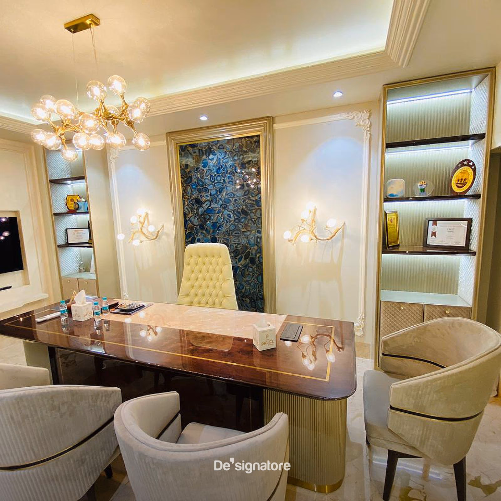 Executive cabin with a wood and gold-trim desk, tufted cream chair, backlit blue agate wall panel and a gold bubble chandelier overhead.