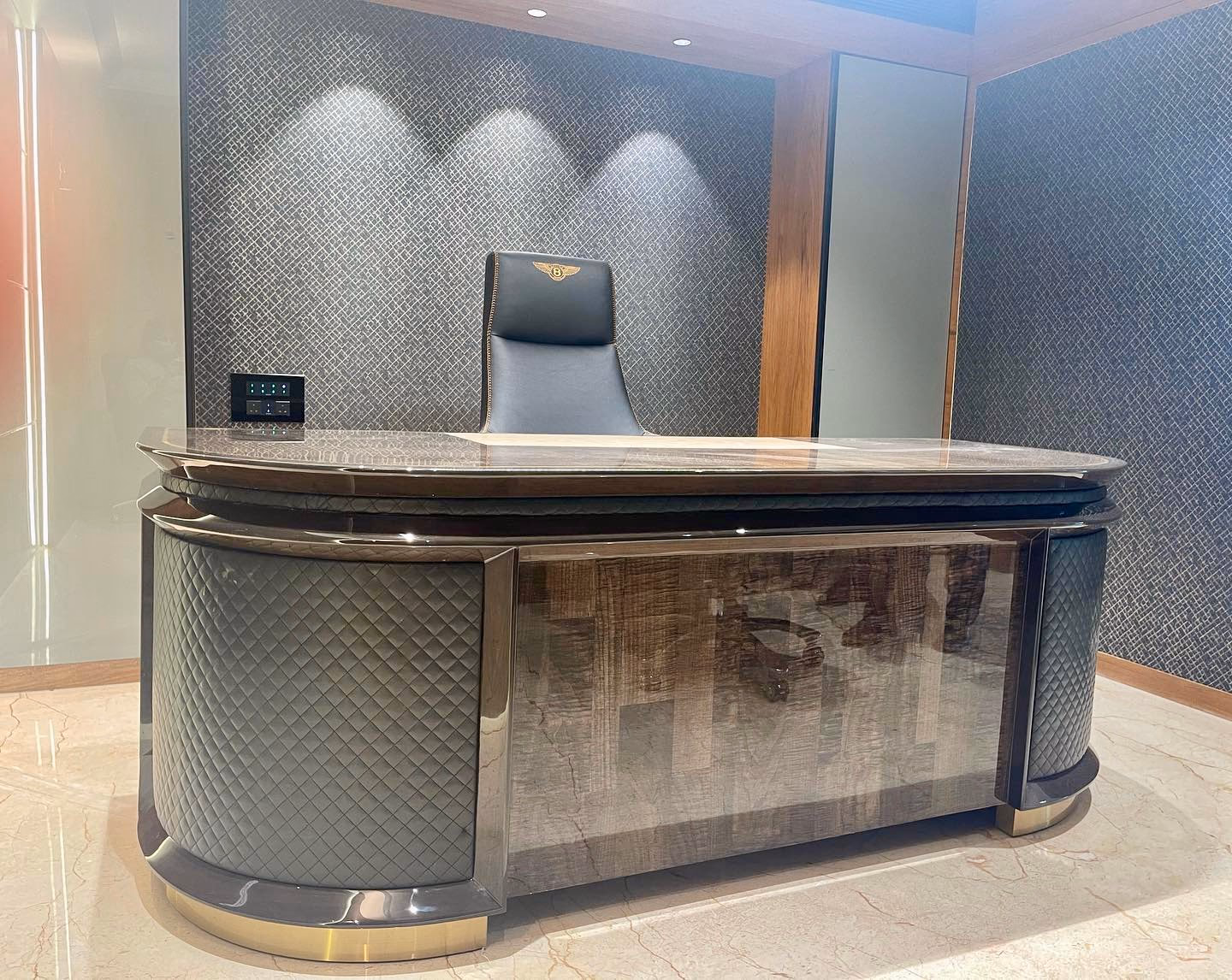Curved executive desk with a dark stone top, grey diamond-quilted leather end pods, a high-gloss burl-wood front panel and polished brass base rings, with a black leather high-back chair behind.