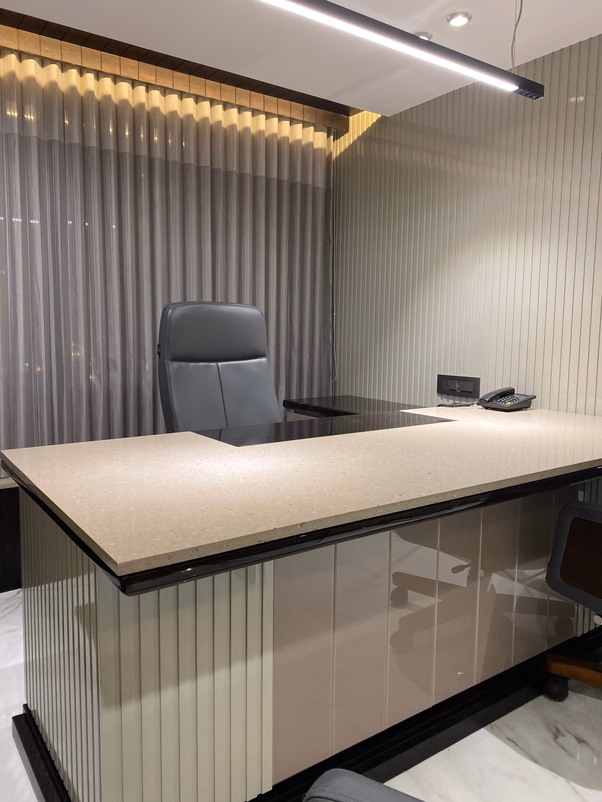 Large L-shaped executive desk with a thick cream stone top over a glossy fluted base, paired with a grey leather chair against sheer curtains.