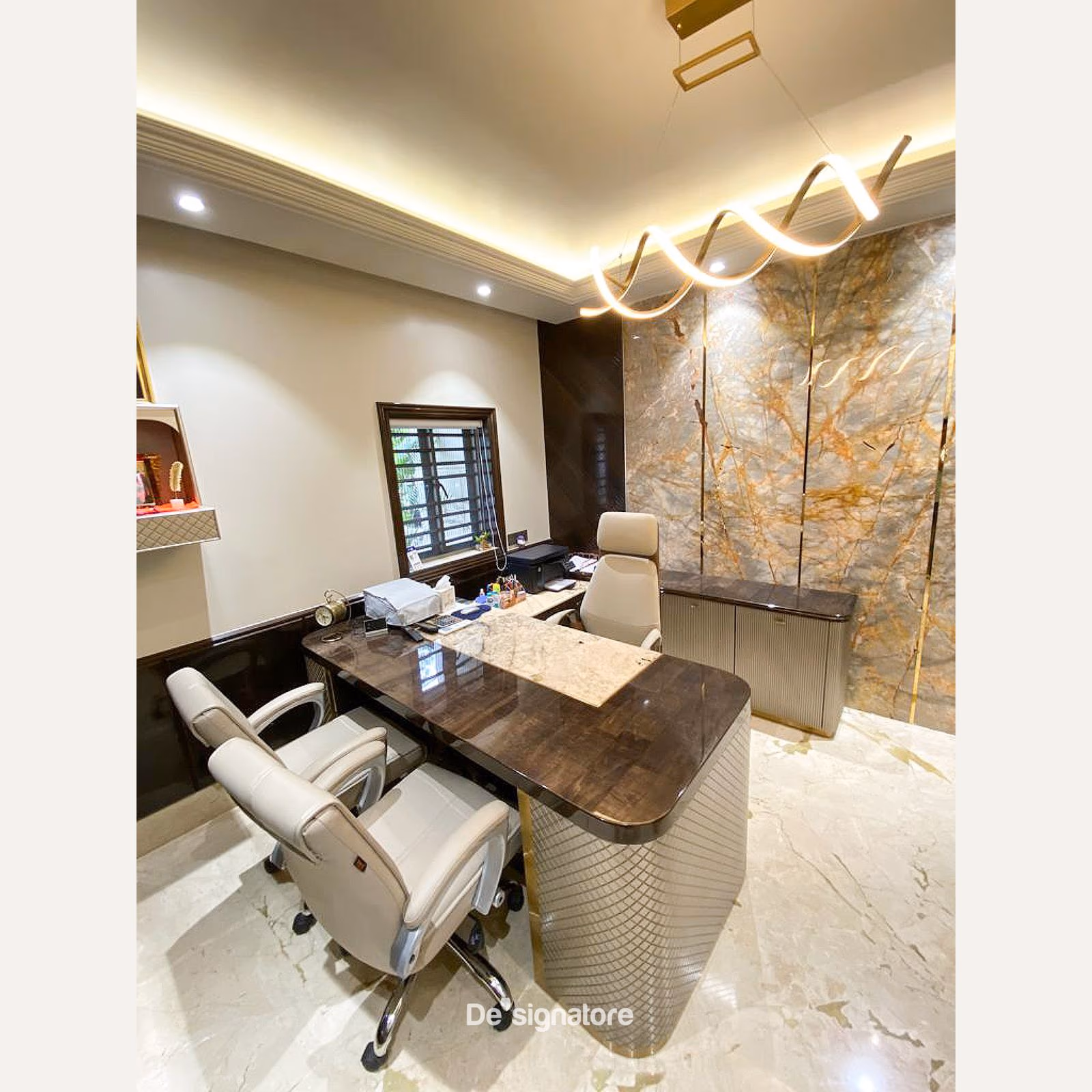 L-shaped executive desk with dark wood and marble top, quilted panel base, twisted gold pendant light and marble accent wall in a corporate cabin.