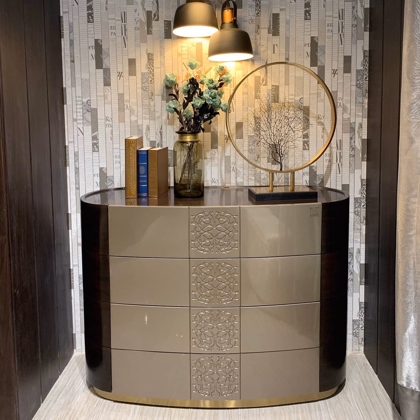 Four-drawer entrance chest in champagne lacquer with a carved medallion centre panel, curved dark rosewood ends and a polished brass plinth, set in a lit alcove.