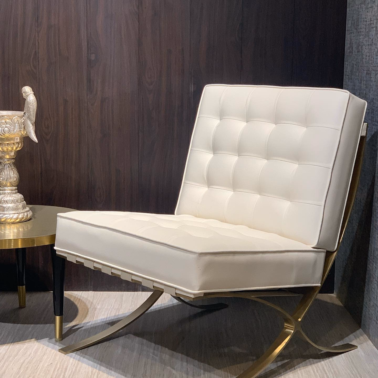 Cream leather lounge chair with a deep square-tufted back and seat cushion, carried on a polished brass X-frame, set against dark walnut panelling.