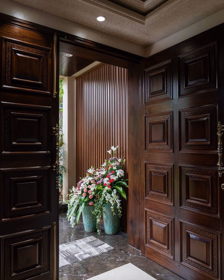 Tall double main door in dark teak with deep raised panels and hand-carved geometric borders, standing open onto a fluted wood entrance lobby with brass handles and marble flooring.