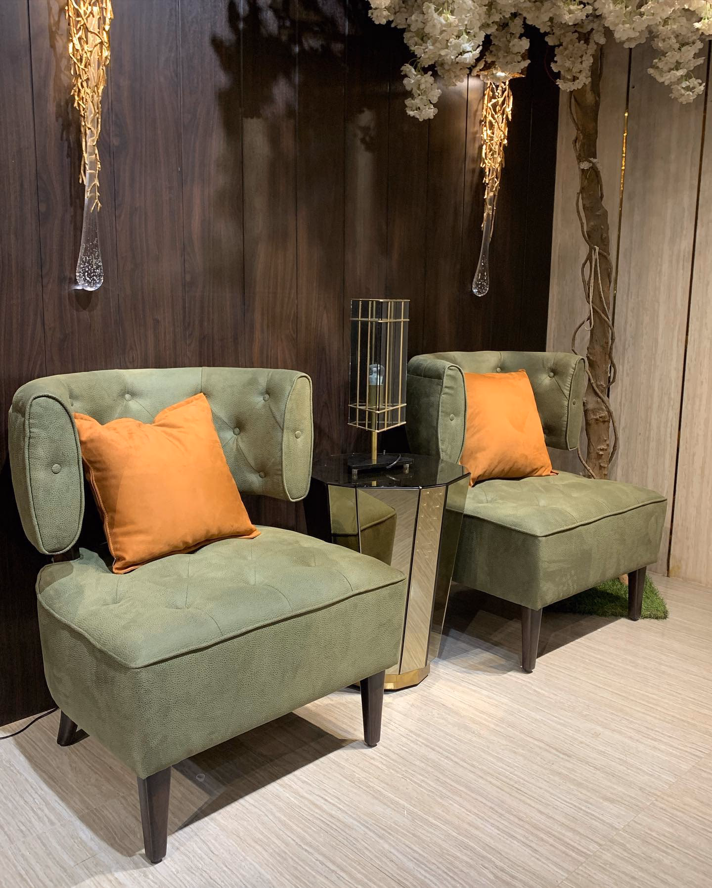 Pair of olive green suede slipper chairs with button-tufted curved half-backs and rust orange cushions, either side of a mirrored drum side table against dark walnut panelling.