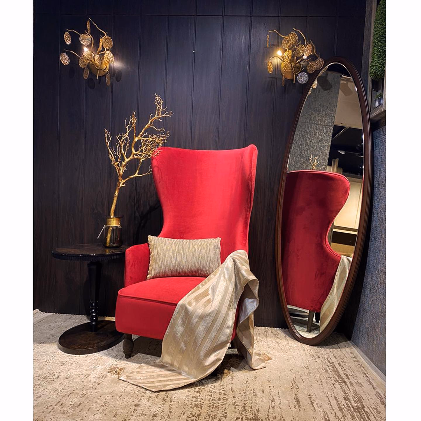Tall red velvet wingback chair with a curved crown, low rolled arms and turned wooden legs, standing beside an oval dark-wood floor mirror against a black panelled wall.