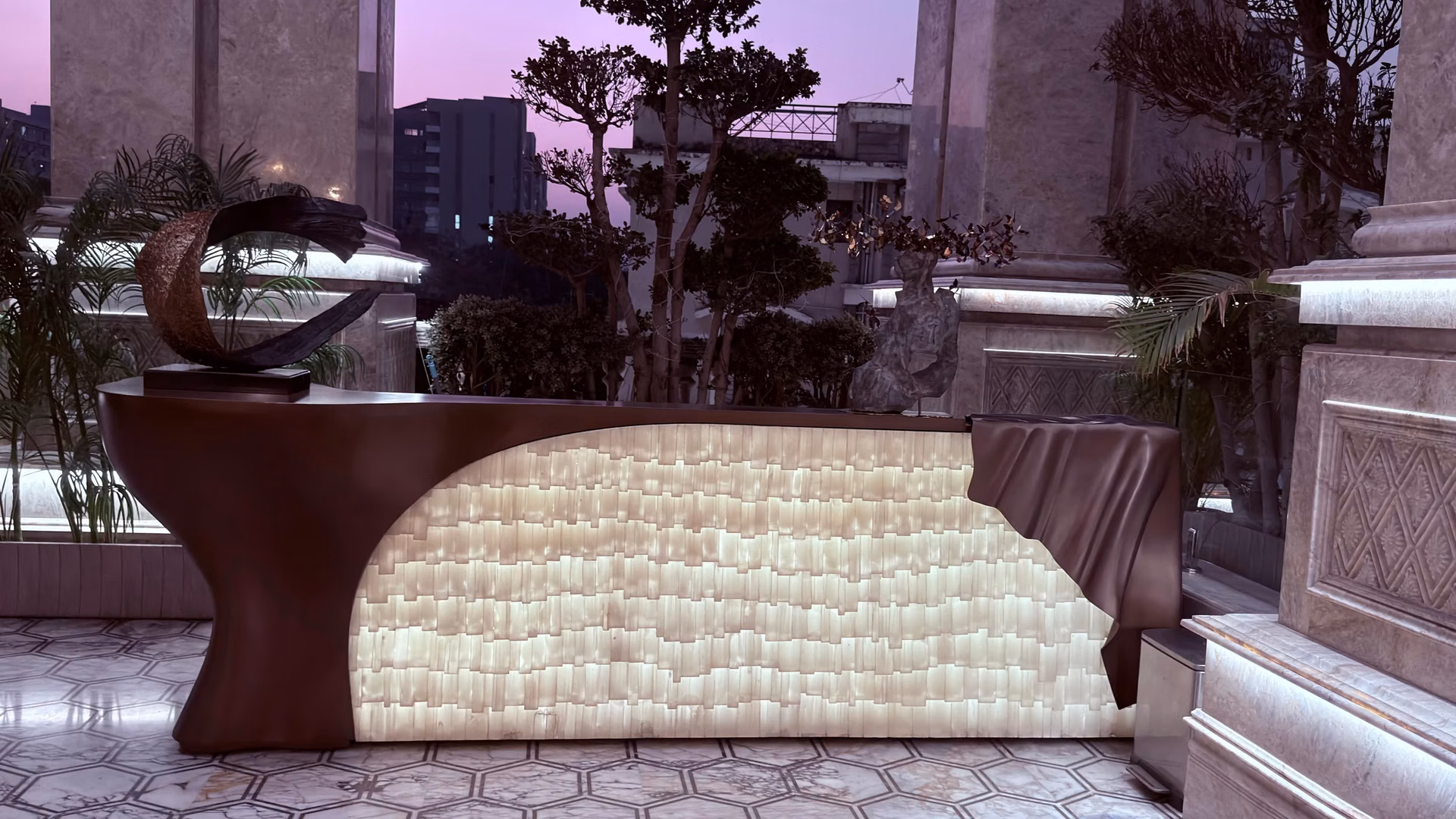 Curved home bar counter with a dark sculpted top and a backlit honey-onyx front that glows warmly, set on a marble terrace at dusk with potted trees.