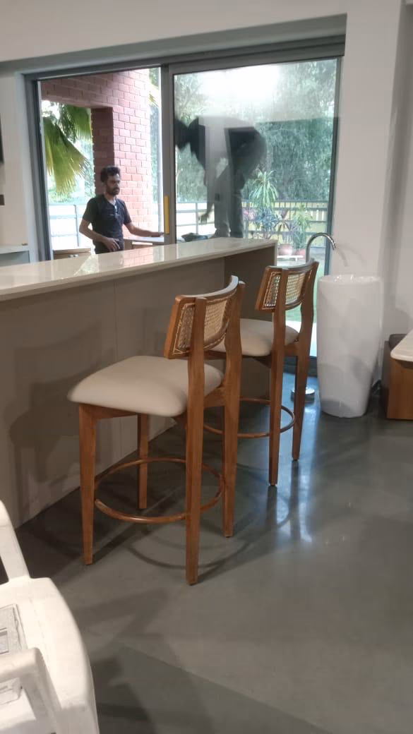 Two tall teak bar stools with woven cane backrests, cream padded seats and round footrest rings, tucked under a stone breakfast counter by a garden window.