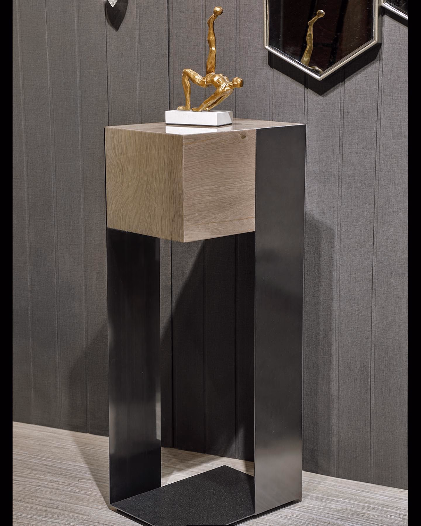 Tall pedestal display table with a pale oak cube top set into a folded black steel frame with an open triangular base, holding a brass figure against grey panelling.