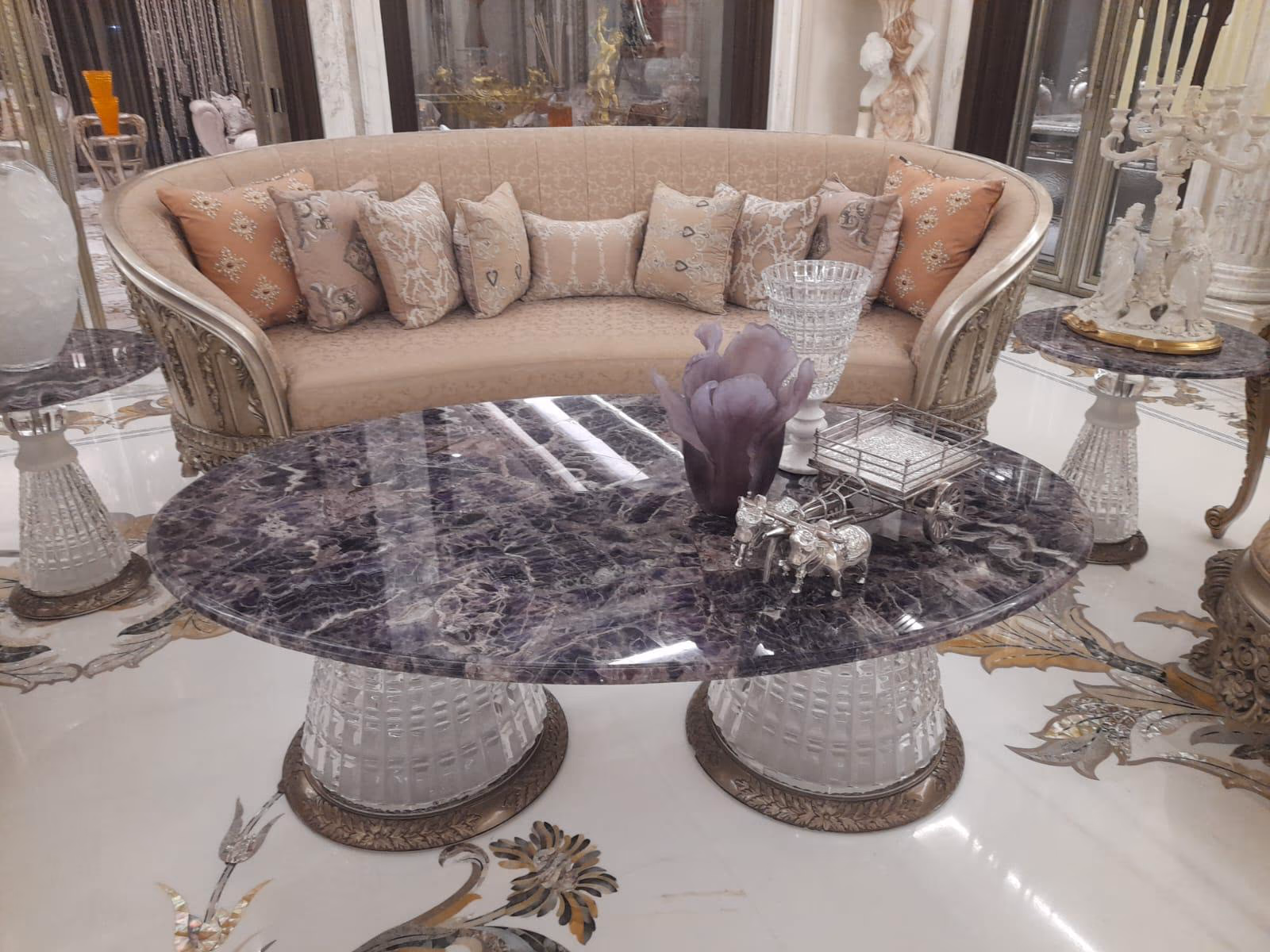 Oval coffee table with a deep purple amethyst stone top resting on two textured crystal drum bases with bronze rims, in front of a curved beige sofa.
