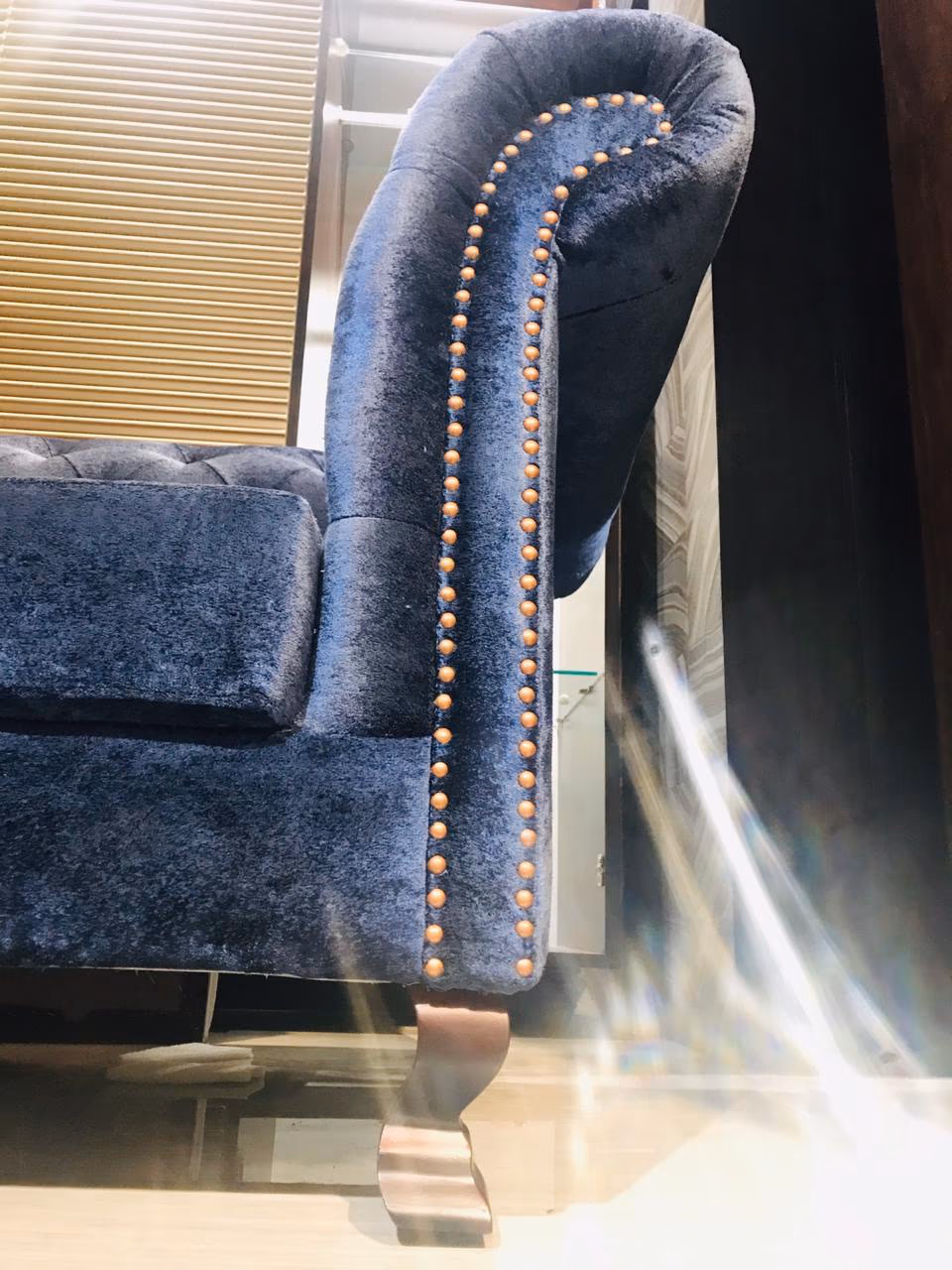 Close-up of the sofa's rolled arm in blue velvet, showing the double row of copper studs curving down to a carved wood foot.