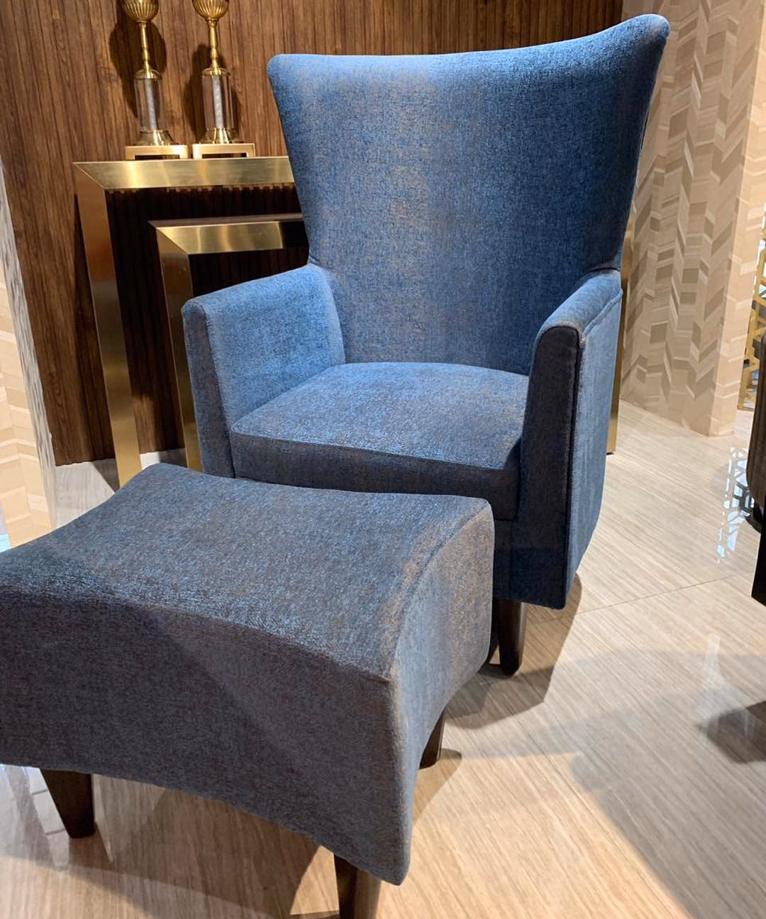 Blue-grey chenille wingback armchair with a curved crown, low arms and dark tapered legs, shown with a matching flared-top footstool ottoman in front of it.