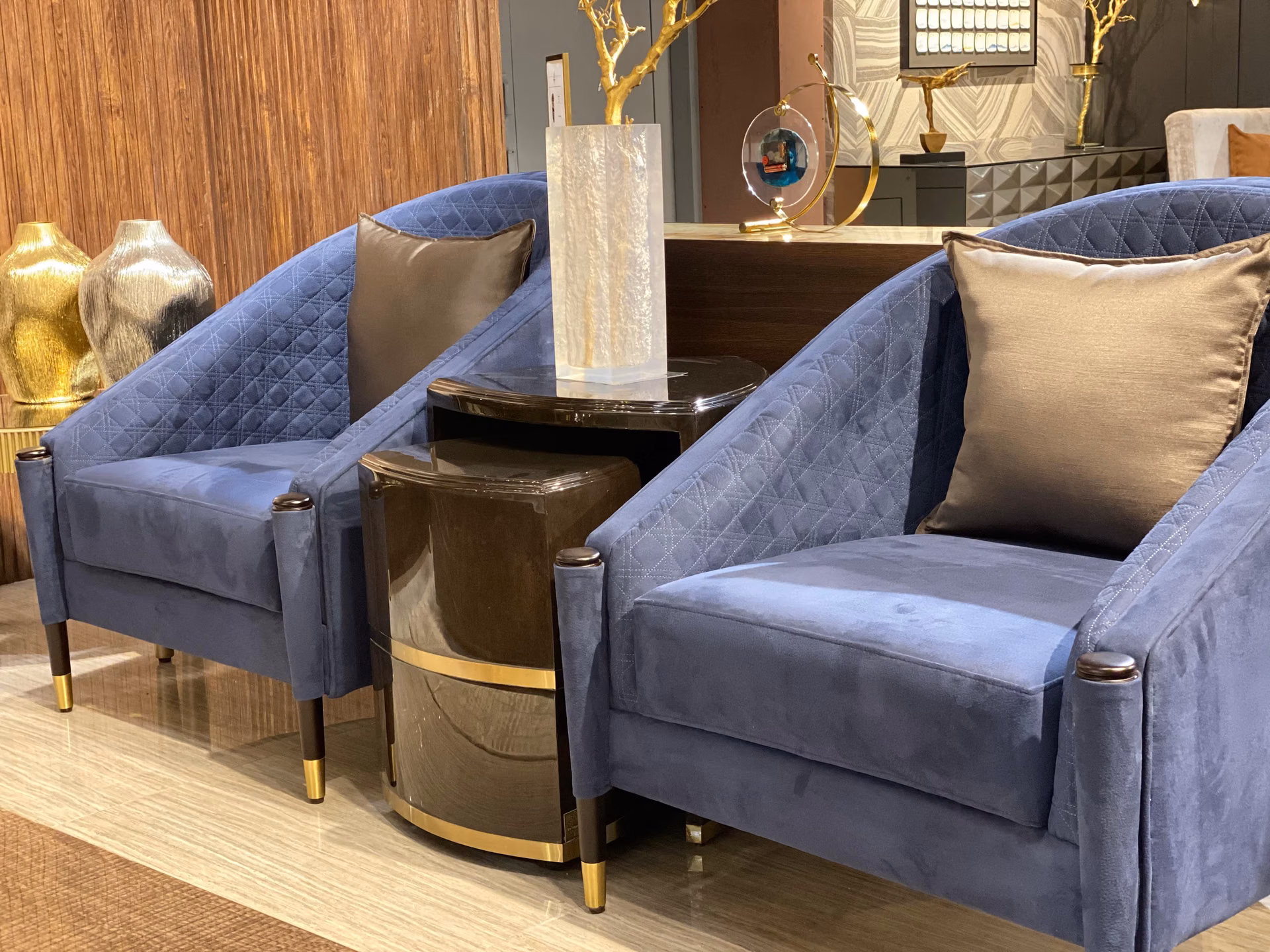Pair of blue velvet armchairs with diamond-quilted backs and brass-capped dark wood legs, set either side of a small round side table against a textured wall.