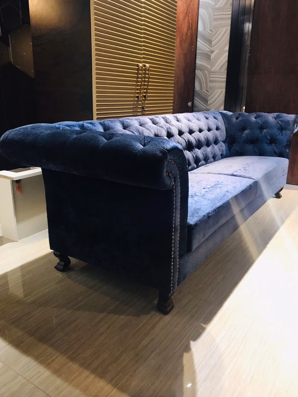 Three-seat Chesterfield sofa in blue crushed velvet with deep button tufting, rolled arms, copper nailhead trim and dark wood bun feet, seen from the side.