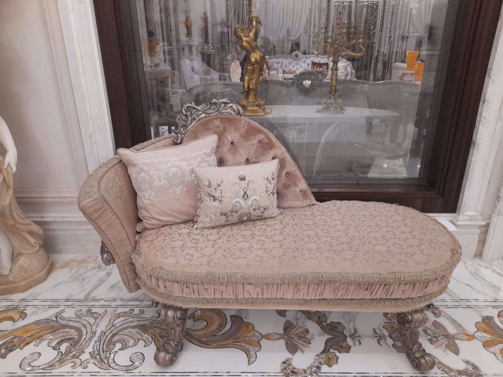 Single-ended blush chaise longue with a tufted velvet backrest, silver-carved crest and frame, fringed skirt and two embroidered cushions on a marble floor.