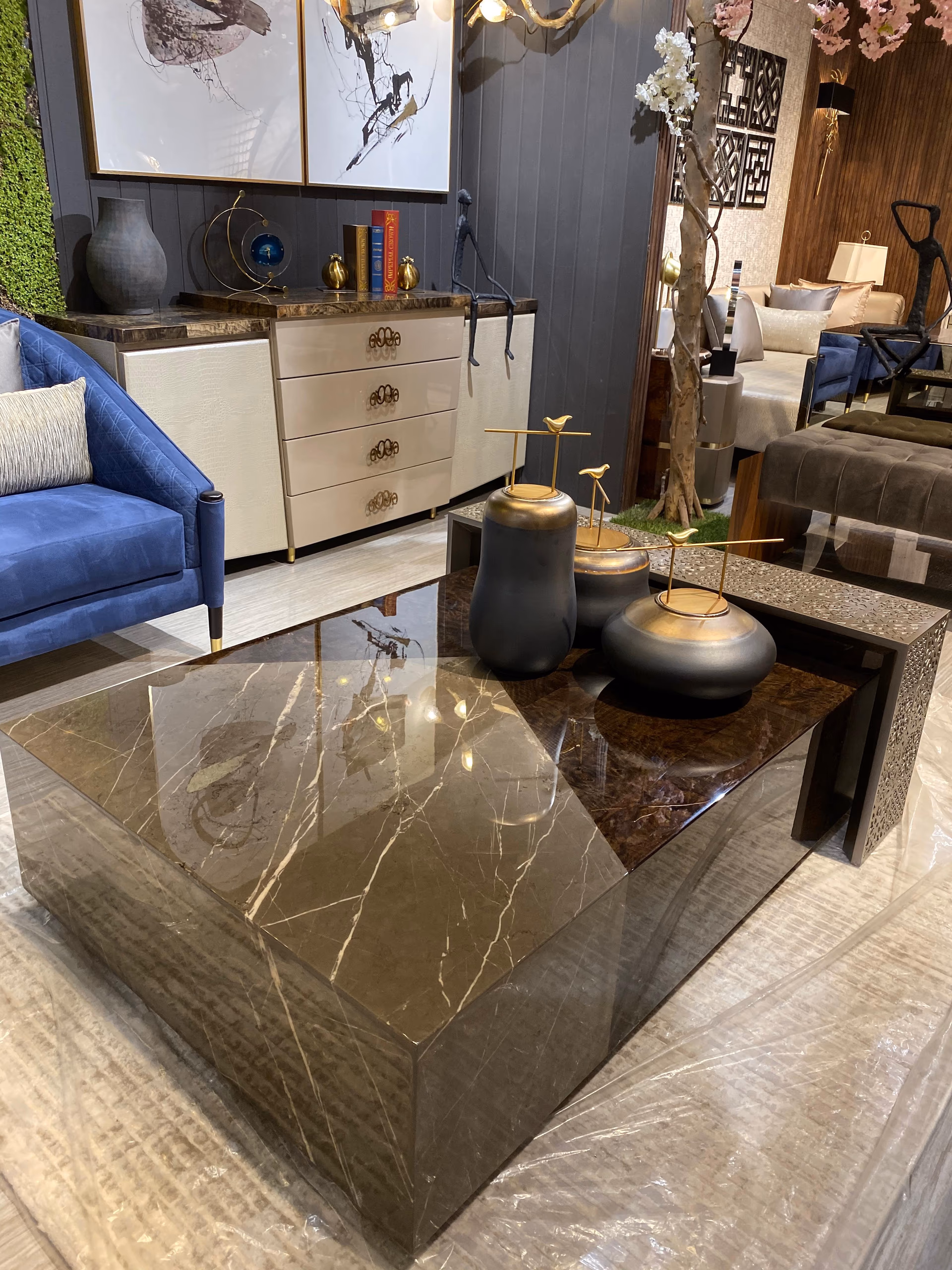 Large square coffee table clad in glossy brown marble with waterfall edges, paired with a smaller pierced-metal nesting table, blue sofa and white sideboard behind.
