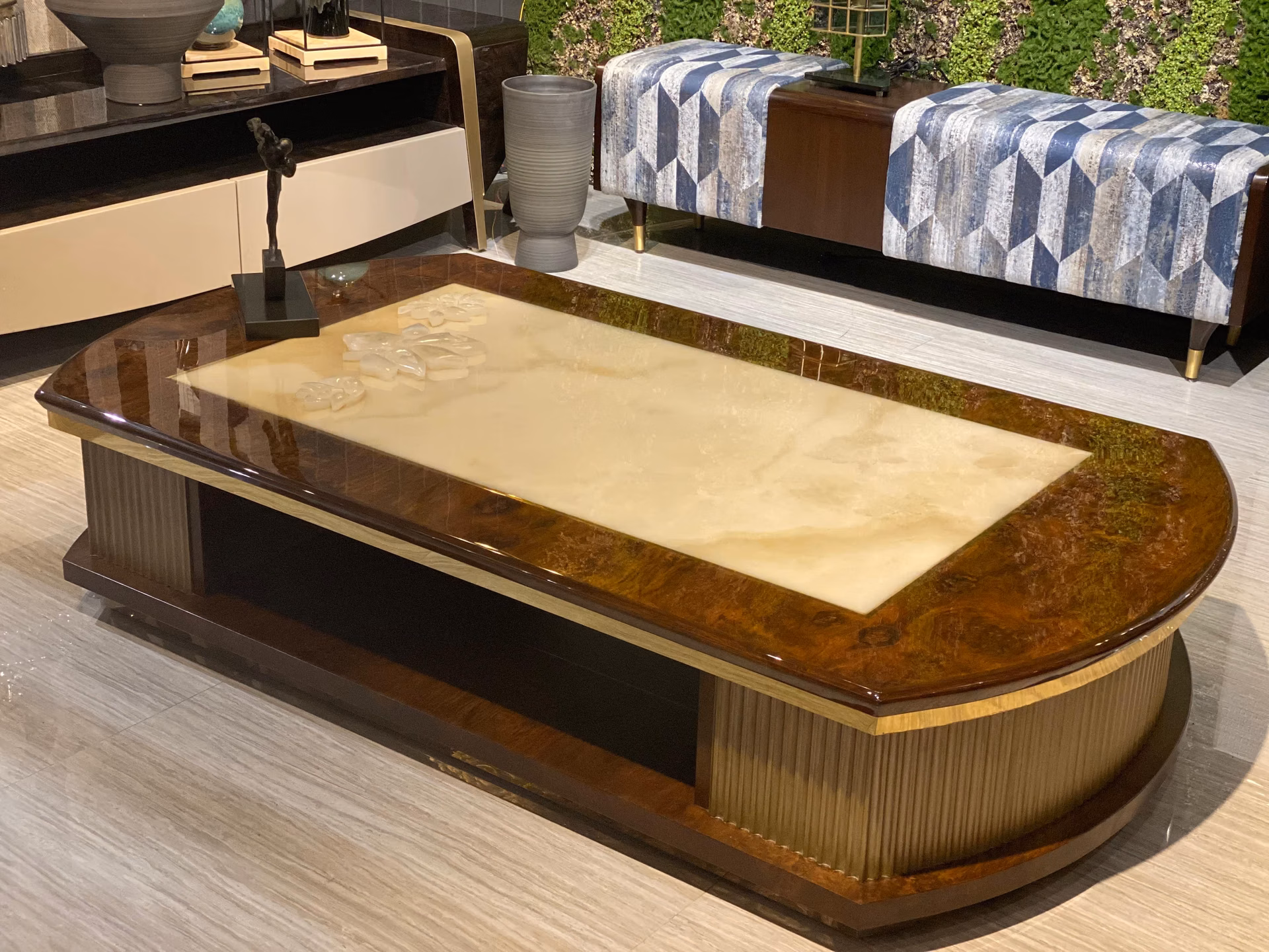 Long oval-ended coffee table with a glossy walnut-burl frame around a carved cream onyx top, on a low metallic fluted band, in a blue-sofa setting.