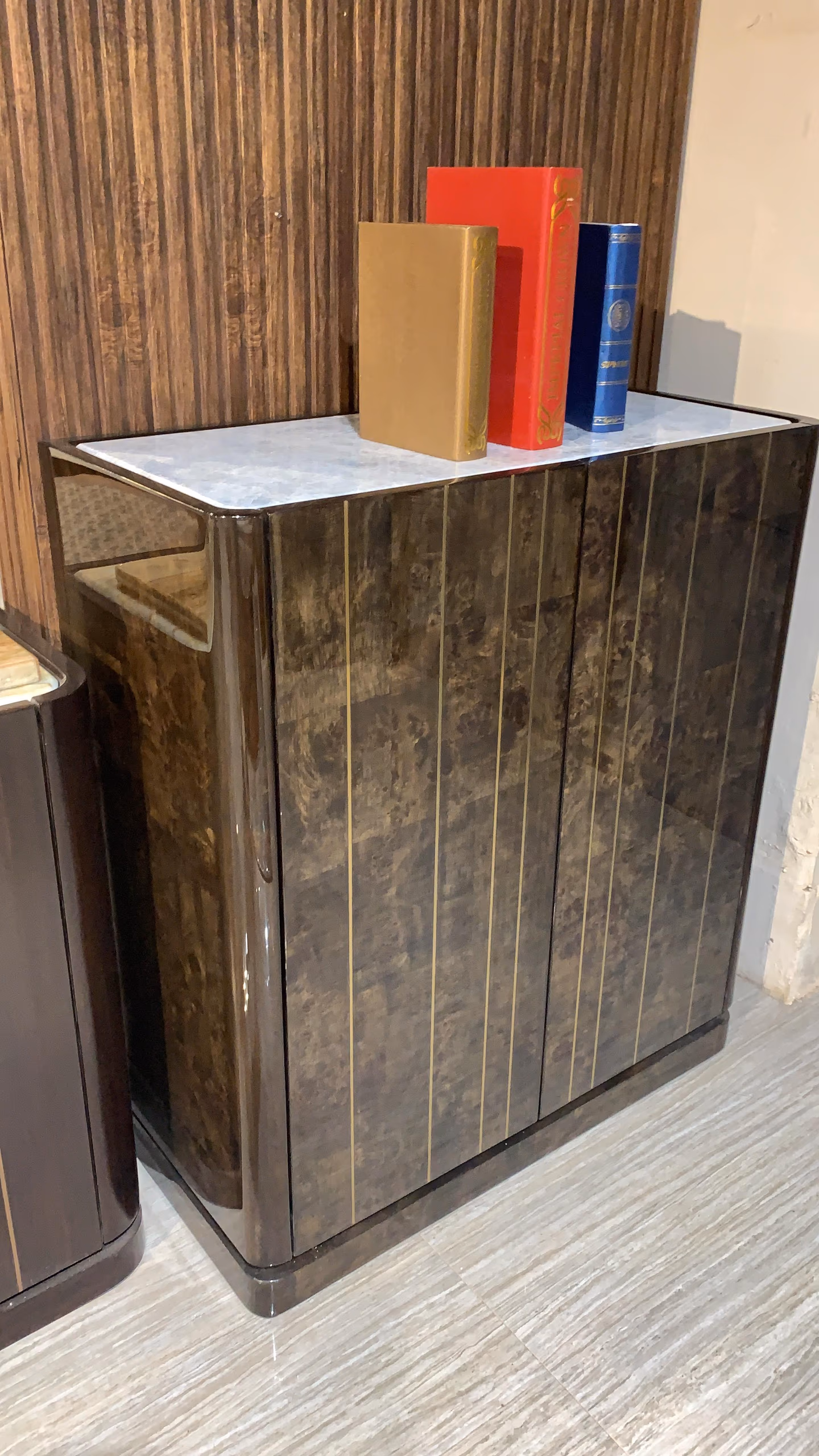 Two-door storage cabinet in high-gloss dark burl walnut with thin brass pinstripe lines and a pale marble top, standing against a showroom wall.