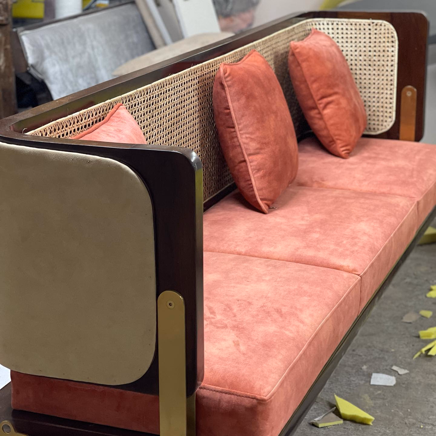 Cane-backed swing seat in blush suede with cream leather outer arms and brass straps, photographed part-built on the workshop floor of the Designatore factory.