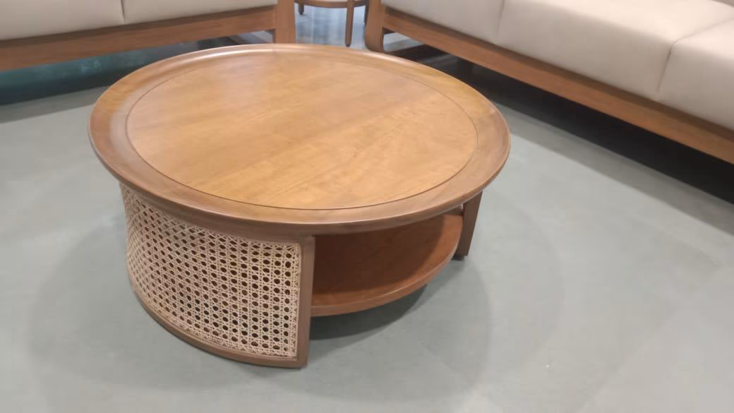Round solid-teak coffee table with a recessed lip top, a lower display shelf and woven cane panels wrapping the drum base, on a grey concrete floor.
