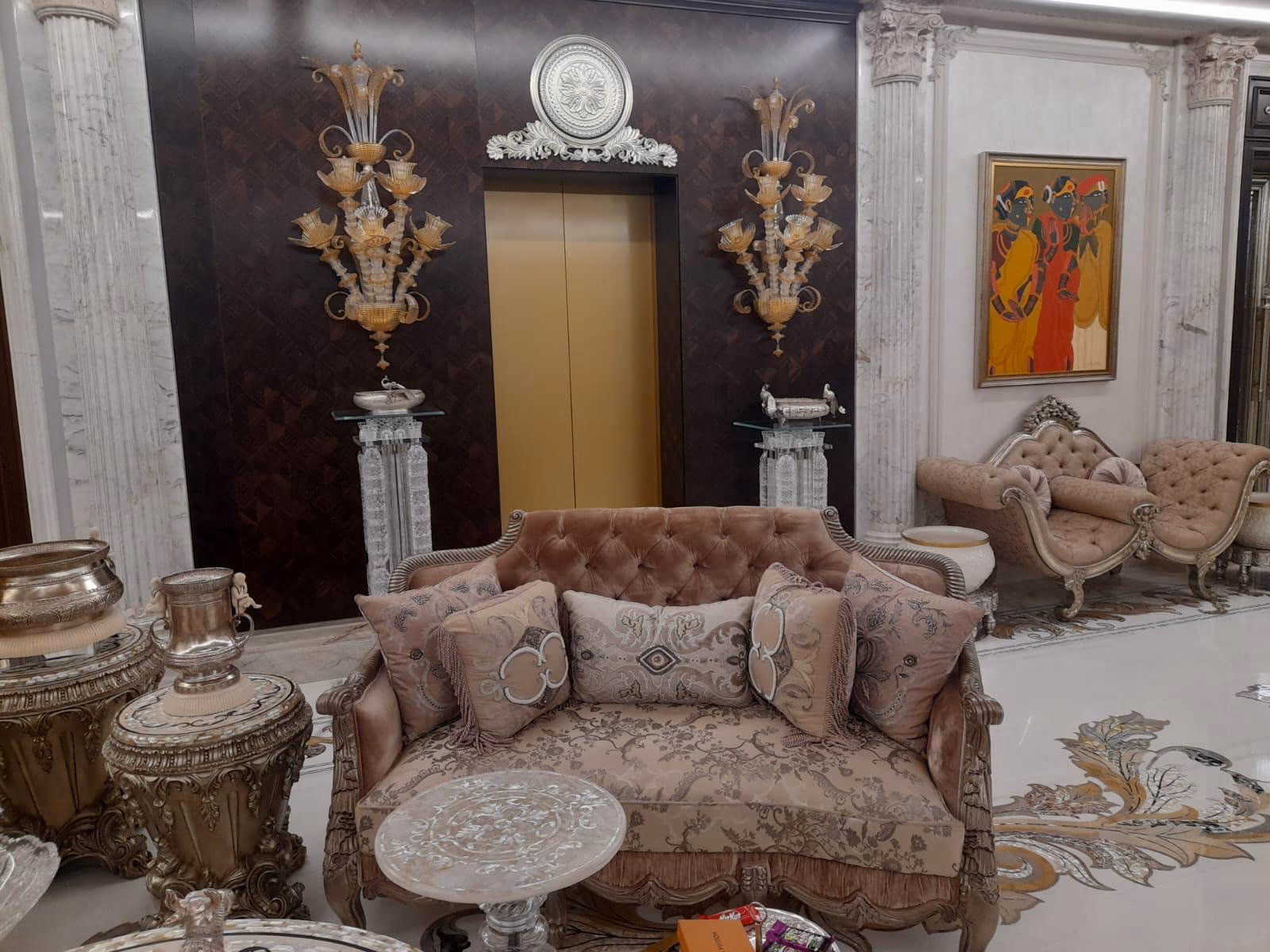 The blush two-seat carved sofa seen at an angle in a marble hall, standing on an inlay floor beside silver urns with a matching chaise in the background.