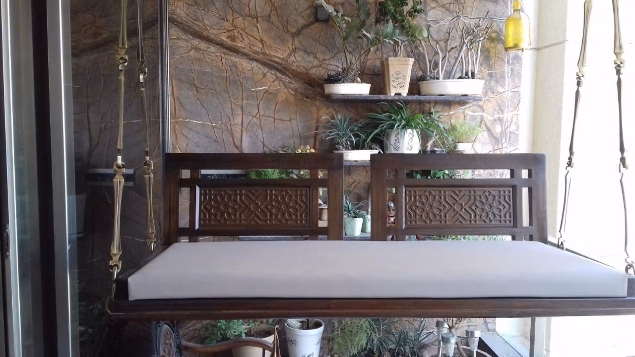 Dark teak indoor swing bench with a carved lattice back panel and a long cream seat cushion, hung on woven straps beside a stone wall and plants.