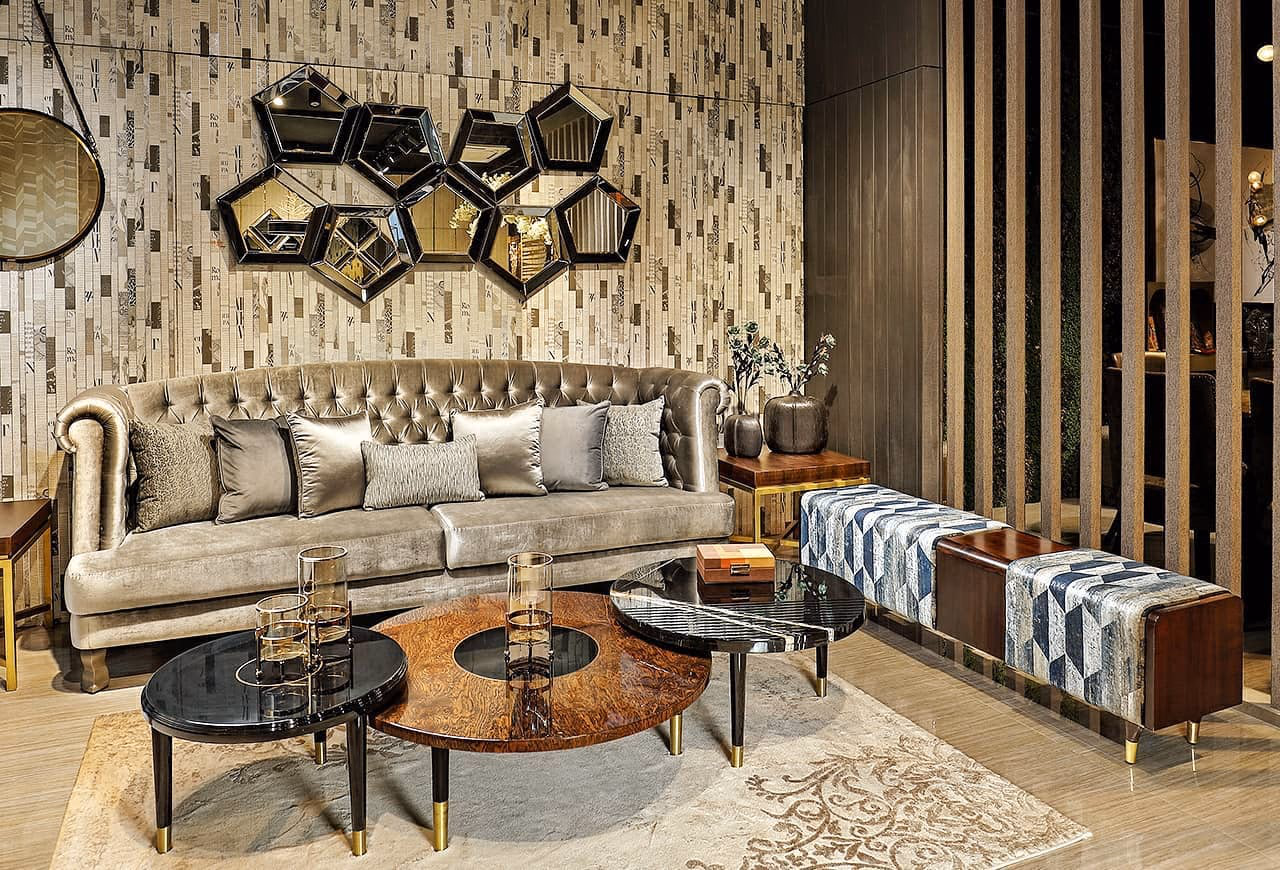 Showroom living room with a champagne velvet deep-buttoned chesterfield sofa, a group of round black marble and burl-wood nesting coffee tables on brass-tipped legs, and a hexagonal mirror cluster on a textured wall.