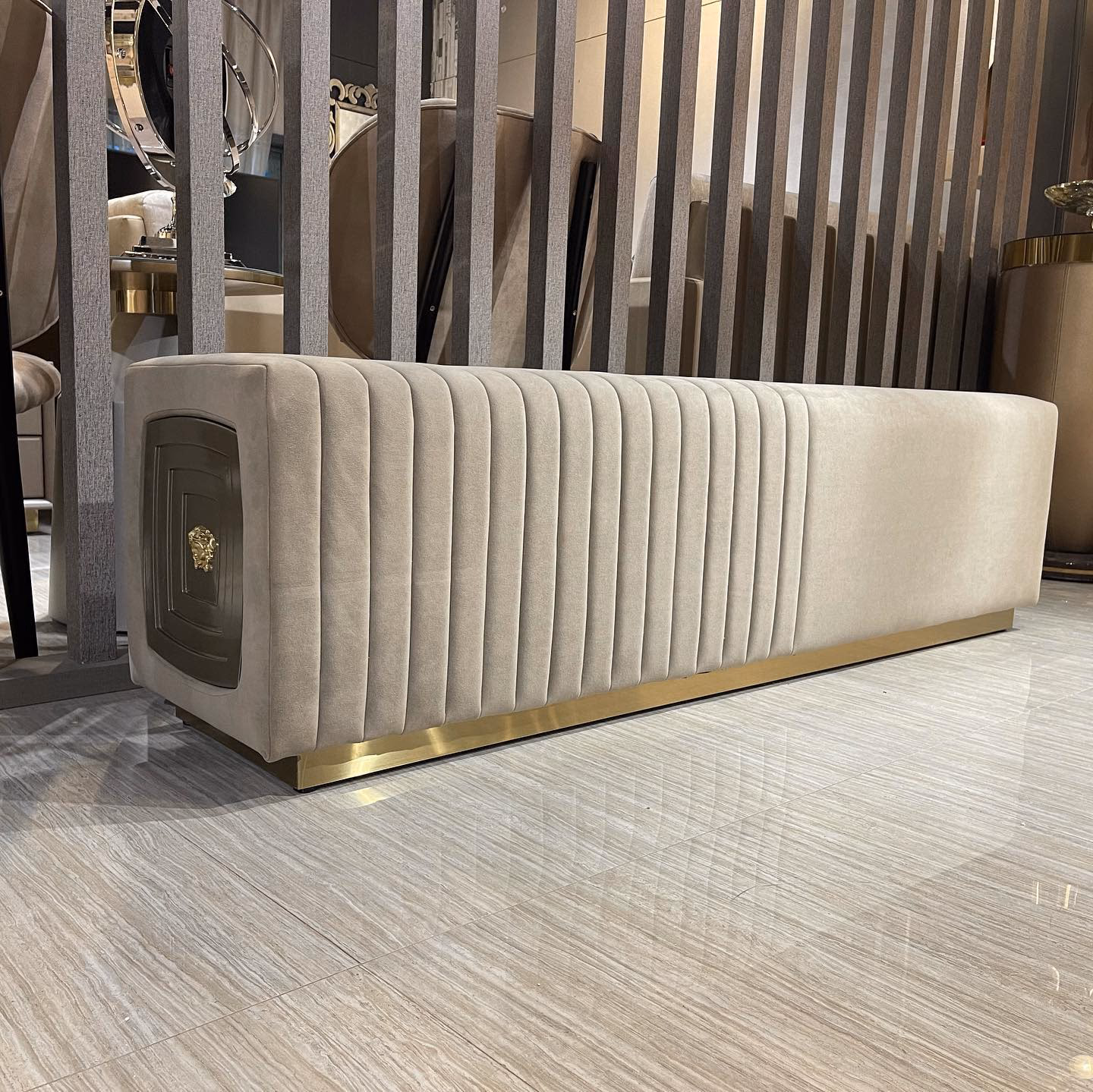 Long cream suede bench with deep vertical channel tufting along the sides, a gold lion-head medallion set into the end panel and a polished brass plinth base.