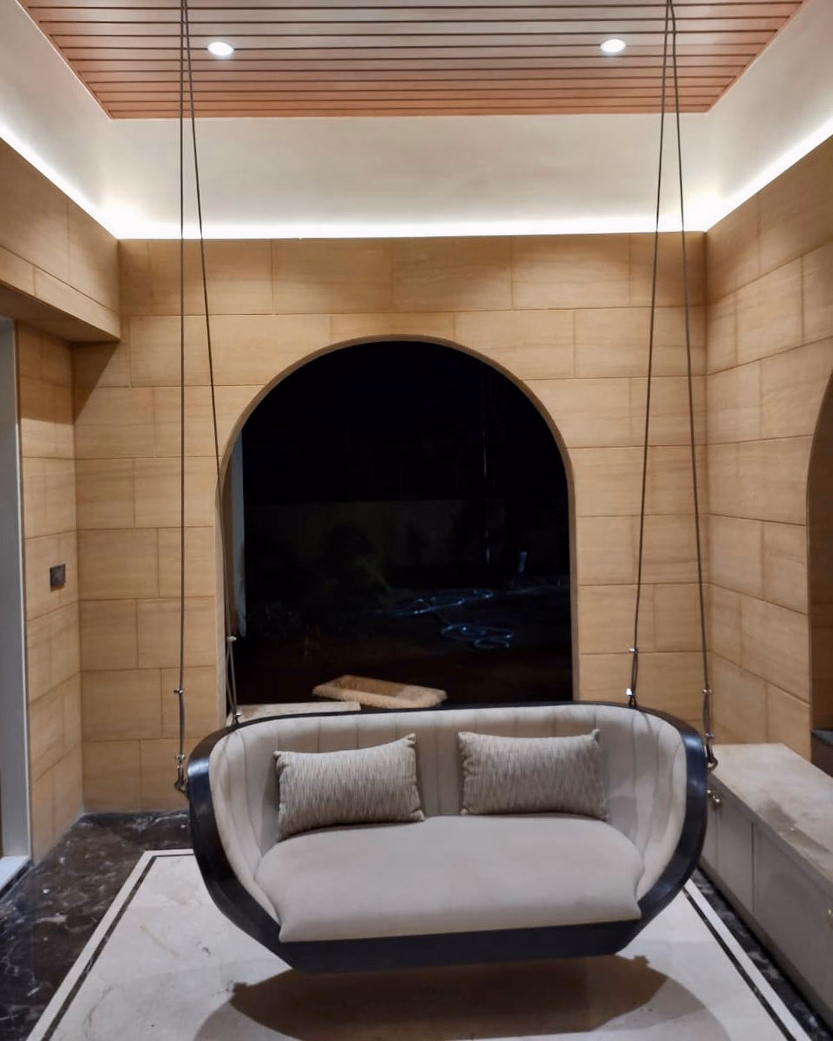 Two-seat hanging swing with a curved dark-lacquer outer shell and a cream upholstered inner seat, suspended on four slim cables in a sandstone-clad alcove.