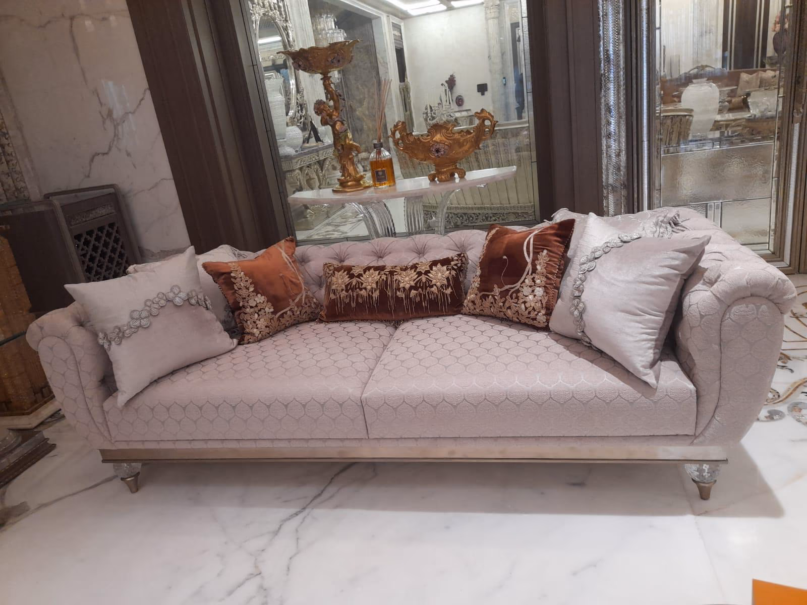 Blush pink tufted three-seat sofa with rolled chesterfield arms, a slim silver base rail and clear crystal front legs on white marble.